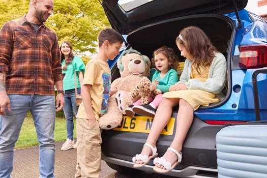 Young children sit in the open boot of a car while parents look on