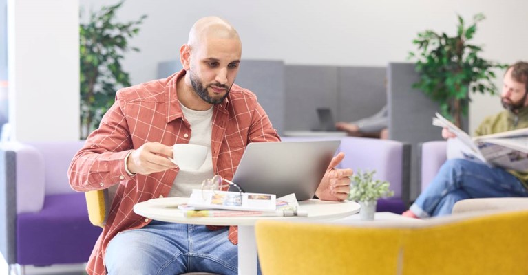 A man holds a hot drink and looks at a laptop